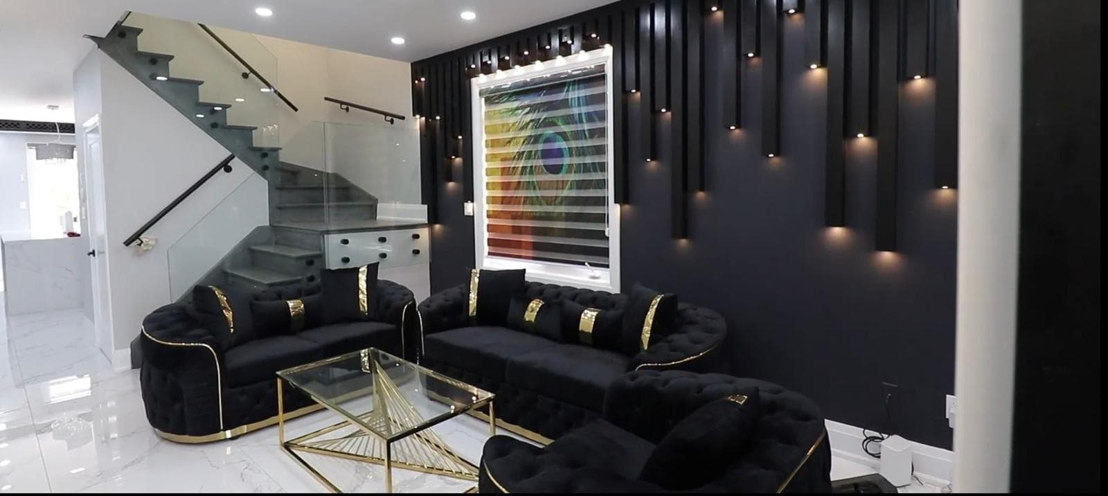 A living-room feature wall in black vertical slats with downlights set between them, beside a glass-balustraded staircase, over a polished marble-effect floor