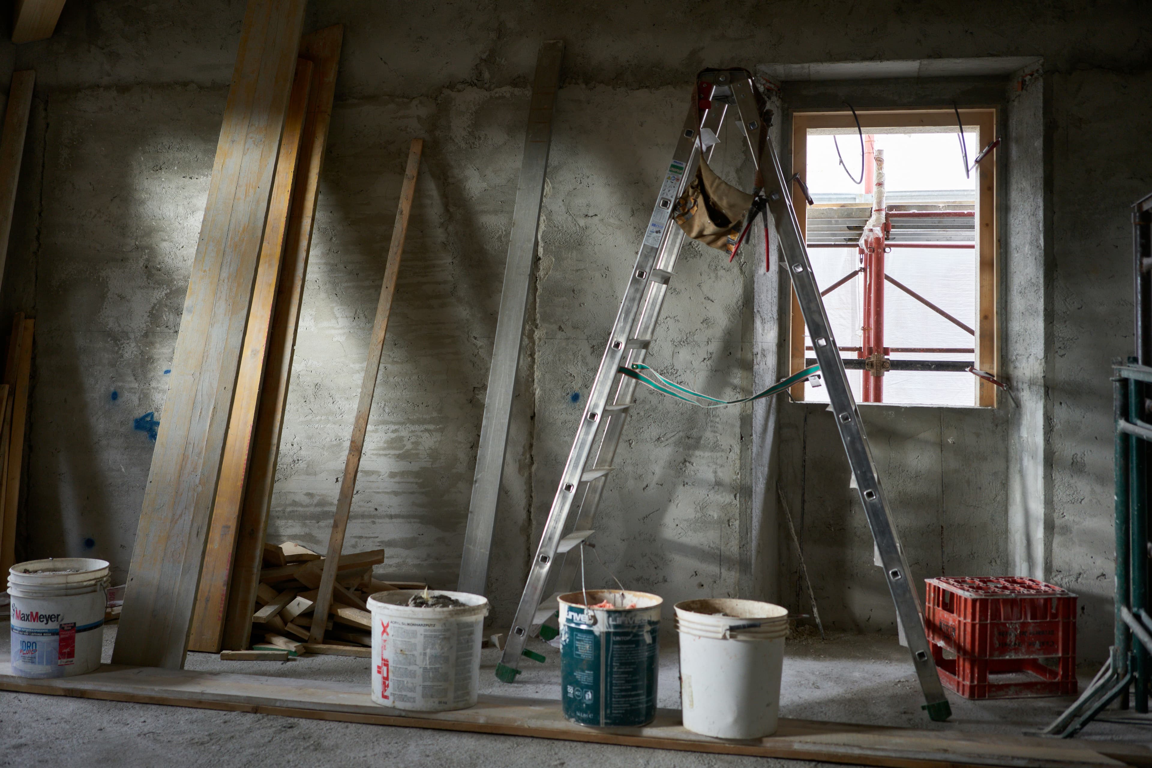 A renovation in progress: bare concrete walls, a step ladder, timber stacked against the wall and paint pails lined up on a plank