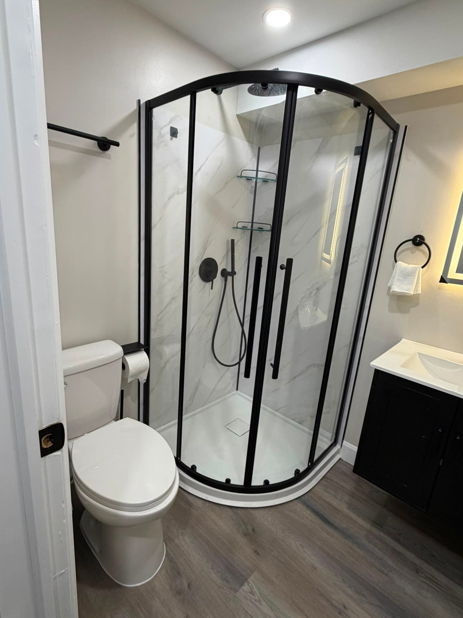 A finished basement bathroom with a quadrant corner shower in marble-effect panelling, black fixtures, a dark vanity and wood-look vinyl plank flooring
