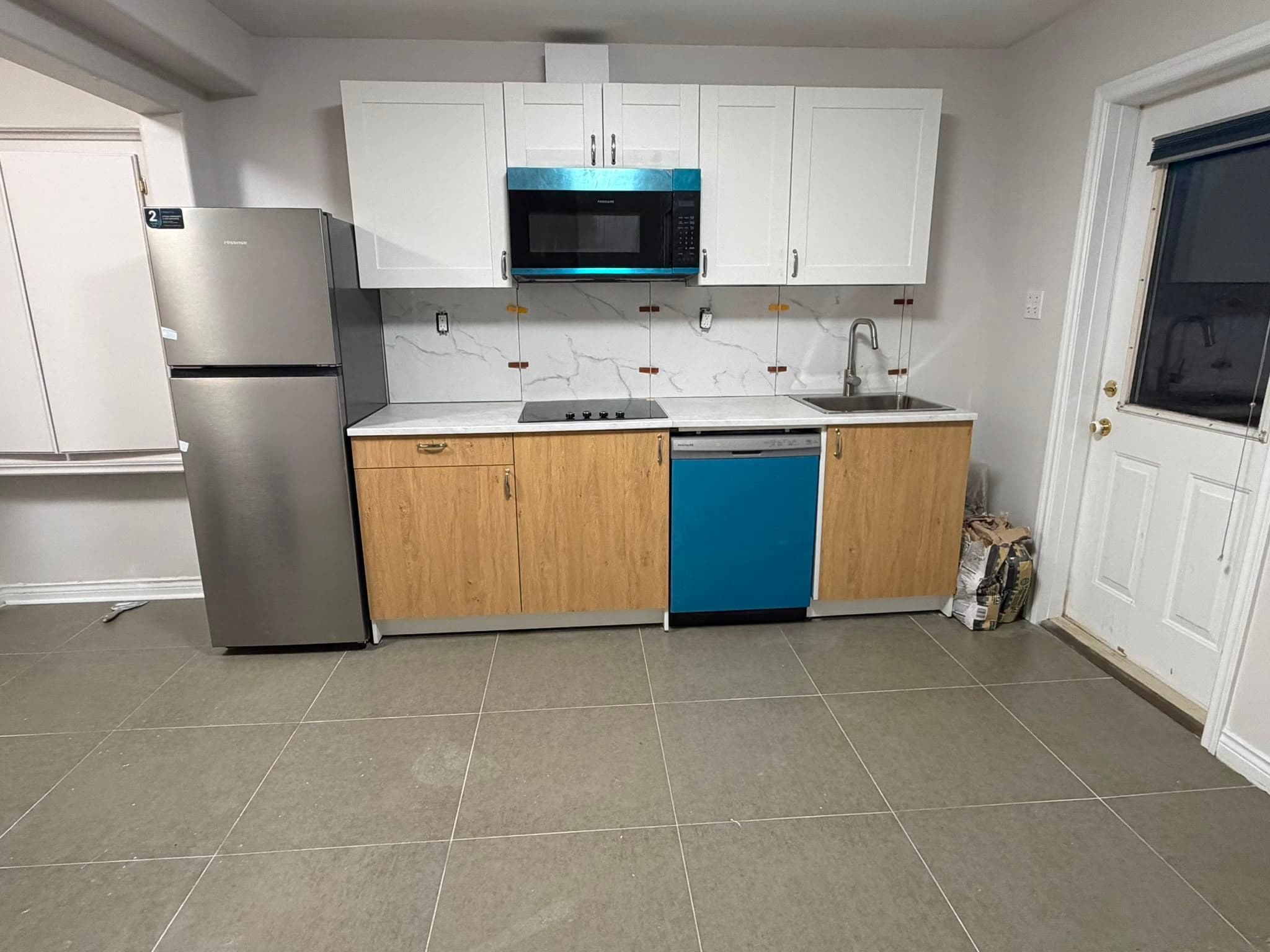 A finished single-run kitchen: white shaker upper cabinets, wood-tone lower cabinets, a white marble-effect counter and matching slab backsplash, drop-in cooktop, undermount sink, over-range microwave and dishwasher, and large-format grey floor tile