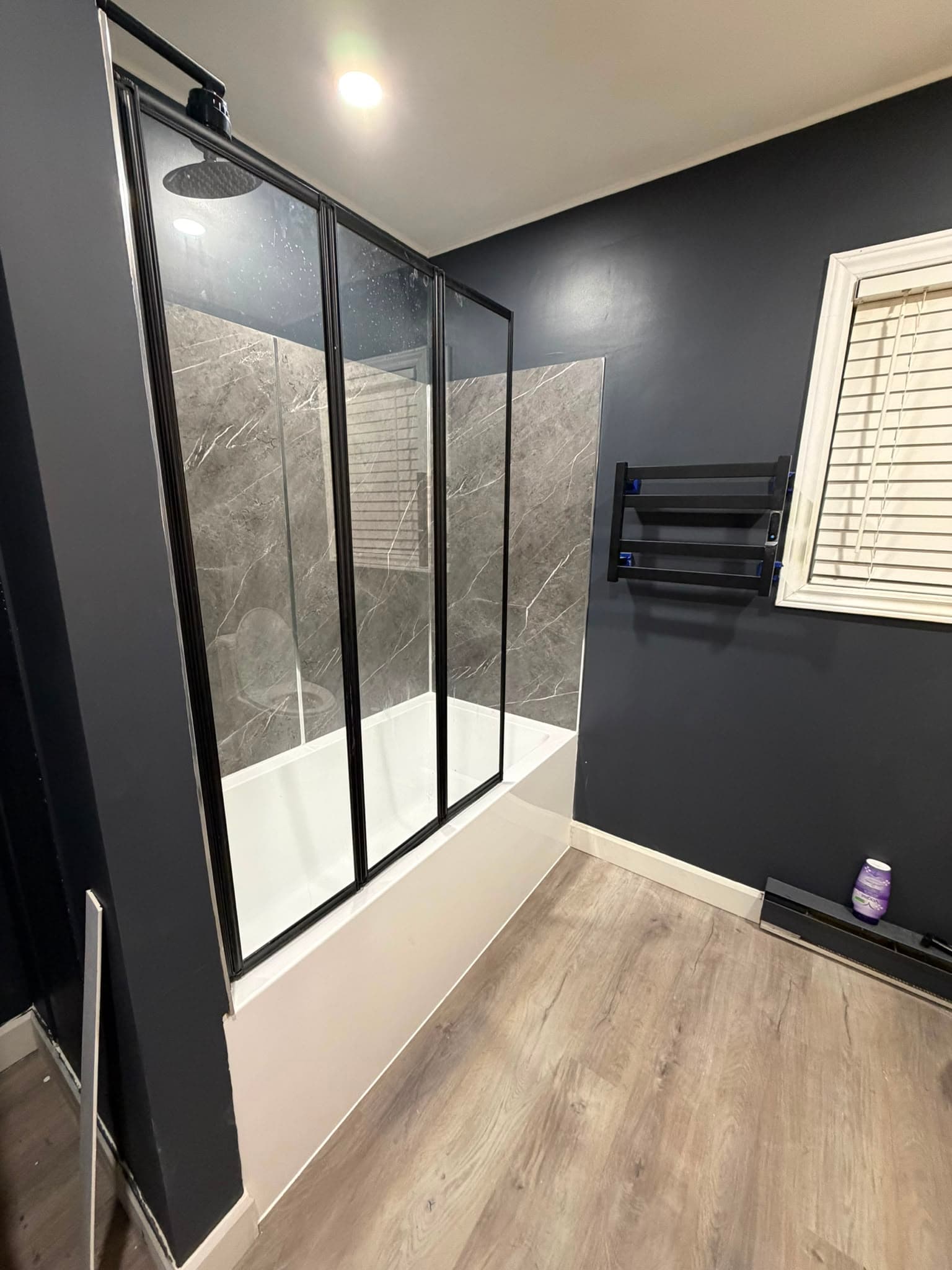 A finished bathroom: white alcove tub with grey marble-effect panelling, a black-framed glass screen, black rain shower head and towel warmer, charcoal walls and wood-look vinyl plank flooring