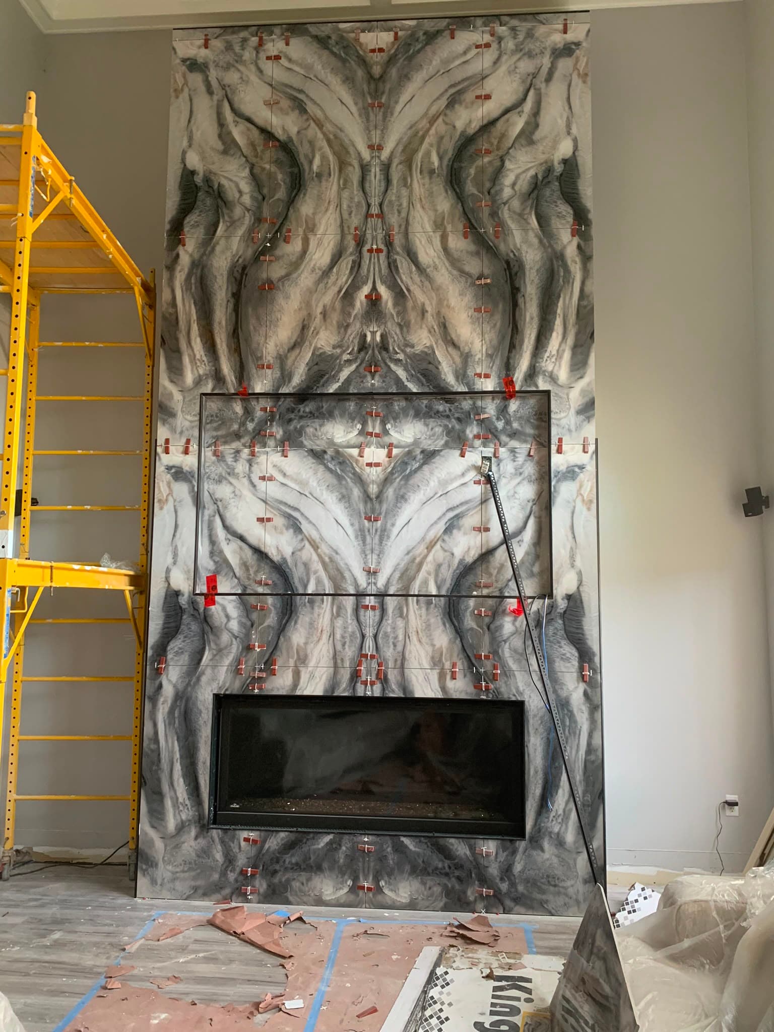 A floor-to-ceiling fireplace wall in book-matched grey and white porcelain slabs, with a linear gas fireplace and a recessed opening above, still carrying tile levelling clips
