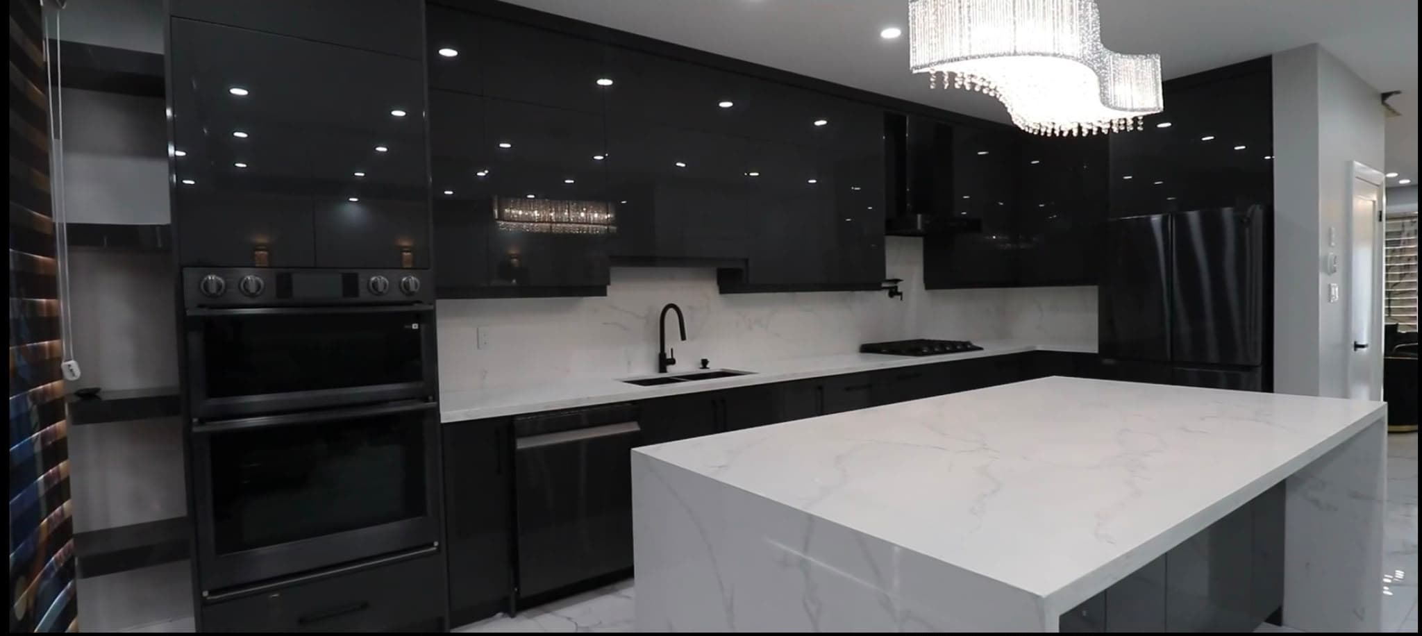 A finished kitchen with high-gloss dark cabinetry, a white marble-effect waterfall island, a tiered crystal chandelier, double wall ovens and a matching slab backsplash