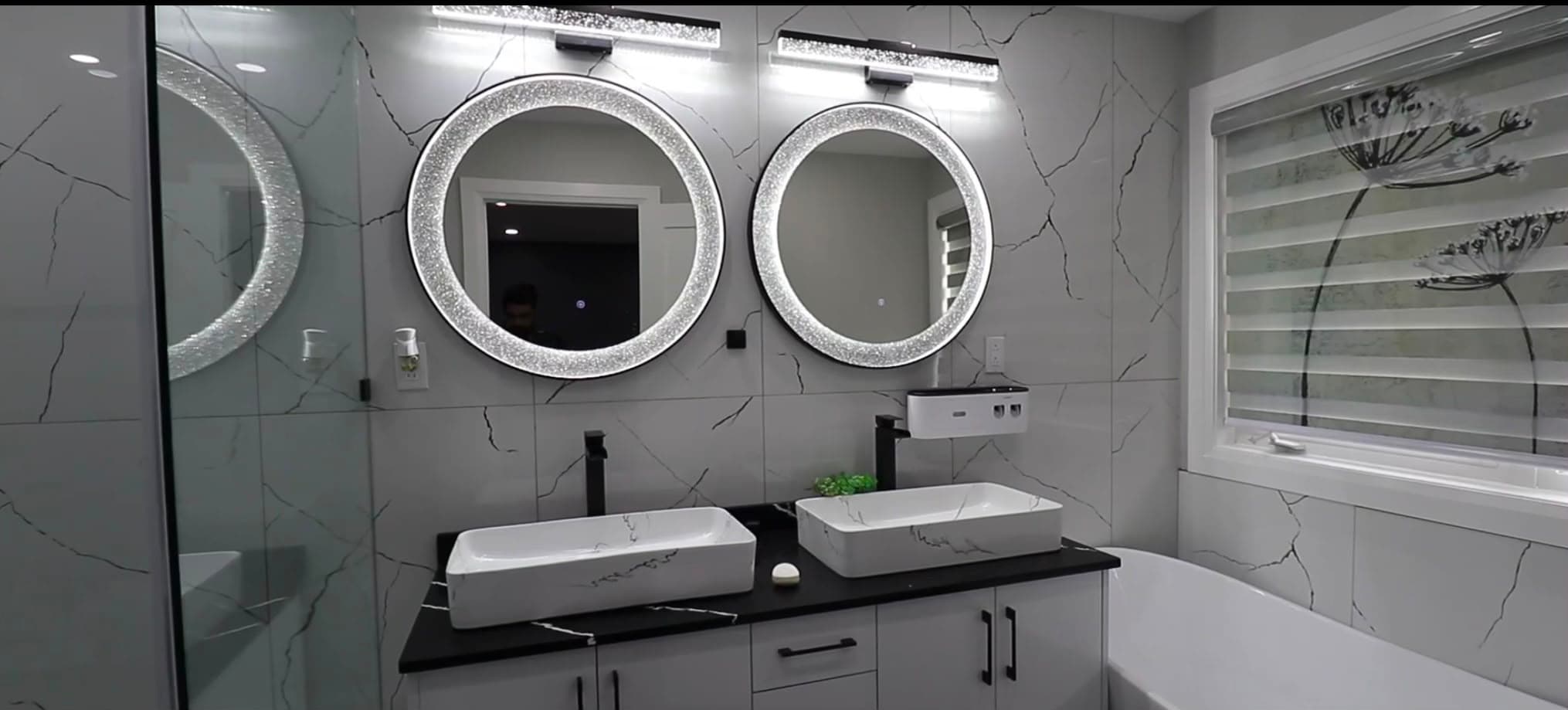 A finished bathroom with two vessel sinks, round lit mirrors and large-format marble-look porcelain