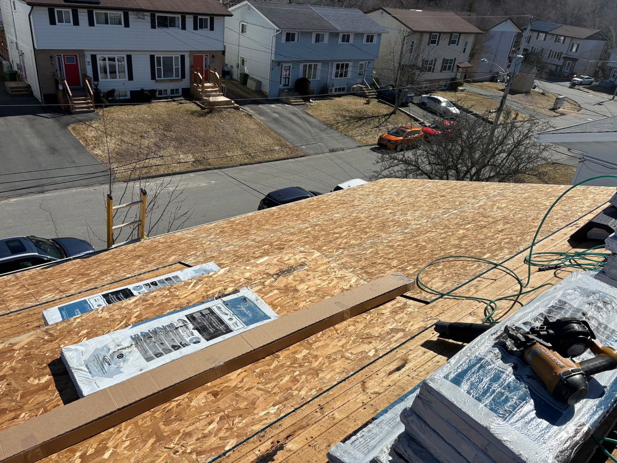 A roof deck sheathed and covered in synthetic underlayment, with shingle bundles staged along the slope and a coil nailer on the ridge